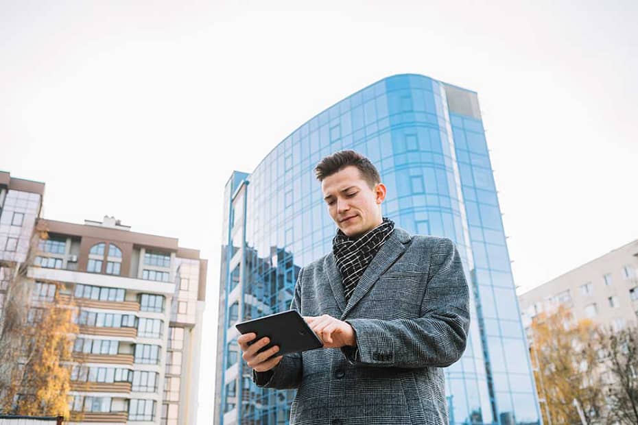Homem de negócios usando um tablet em frente a edifícios modernos, representando a facilidade de gerenciamento proporcionada por um aplicativo para condomínio.