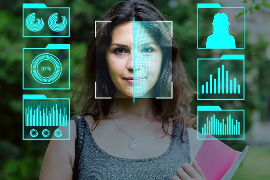 Mapeamento Facial para Condomíio - Mulher em frente a uma tela de reconhecimento facial, com gráficos digitais flutuando ao redor de seu rosto. A interface exibe vários ícones de dados, incluindo gráficos de barras, um indicador de porcentagem e uma silhueta, representando um processo avançado de identificação biométrica. O fundo sugere um ambiente externo com vegetação, contrastando com a tecnologia de ponta exibida na imagem.