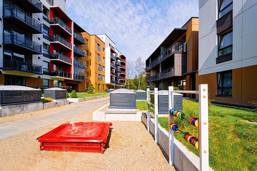 Esta imagem mostra um complexo residencial moderno, composto por vários prédios de apartamentos de diferentes cores e designs arquitetônicos. No centro do complexo, há um espaço ao ar livre bem cuidado com áreas gramadas, caminhos pavimentados e áreas de recreação infantil, incluindo um grande ábaco colorido e uma estrutura vermelha, possivelmente uma caixa de areia coberta. As fachadas dos prédios apresentam varandas com grades metálicas, oferecendo aos moradores vistas para o pátio central. O céu está parcialmente nublado, criando uma atmosfera tranquila e convidativa para os residentes desfrutarem do espaço ao ar livre.